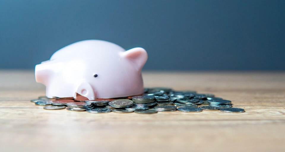 A broken pink piggy bank spilling over with a pile of coins on a wooden table