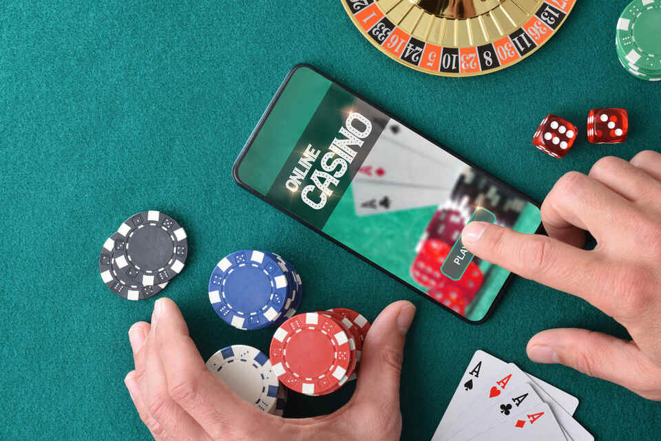 Online casino player entering app to play with hand pressing on smartphone screen on green felt gaming table with betting chips and casino game objects